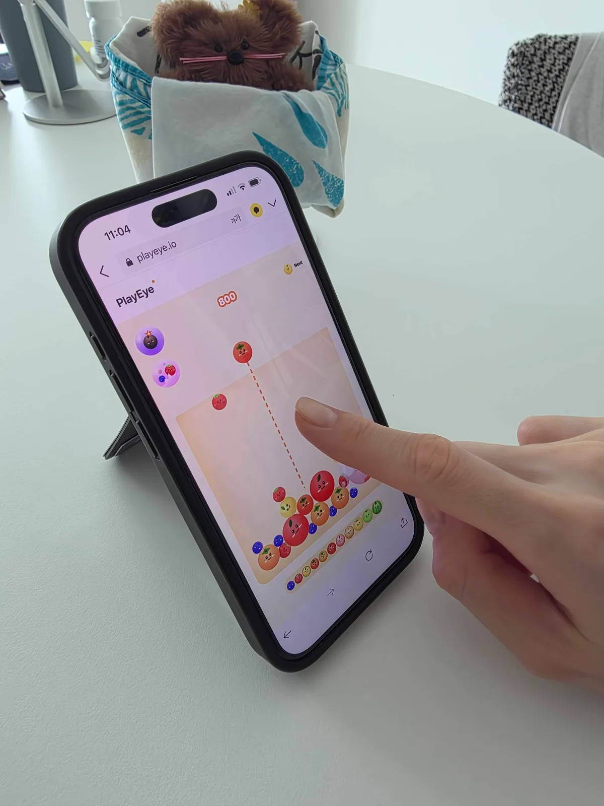 A finger lining up a tomato drop in Watermelon Game at 800 points, with the dotted aim line visible on the iPhone screen